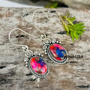 May include: A pair of silver earrings with oval-shaped, multi-colored stones. The stones feature a vibrant mix of pink, blue, and gold hues. The earrings have a decorative border and a hook closure.