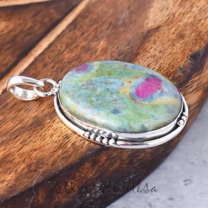 Puede incluir: Un colgante de plata con una piedra preciosa ovalada grande de color verde y rosa con un aro de plata. La piedra preciosa tiene un patrón moteado de verde, rosa y blanco.