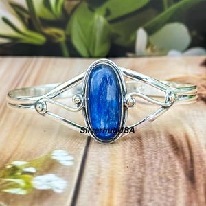 May include: A silver bangle bracelet with a blue kyanite gemstone set in a delicate, ornate design. The bracelet features a curved, open-ended design with a central oval-shaped gemstone.