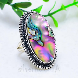 May include: Oval-shaped sterling silver ring featuring an iridescent abalone shell. The shell displays a vibrant array of colors, including pink, green, and blue. The ring has a detailed, beaded border.