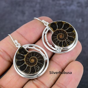 May include: A pair of silver earrings featuring a fossilized ammonite design. The earrings have a circular frame with small decorative beads and a hook closure. The ammonite fossils display a spiral pattern in shades of brown and tan.