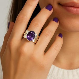 Può includere: Un anello d'argento con una grande gemma di ametista ovale. L'anello ha una fascia larga con un design semplice e moderno. Le unghie della persona sono dipinte di una tonalità viola abbinata. L'anello è un gioiello.