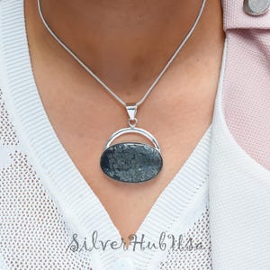 Könnte beinhalten: Eine silberne Kettenkette mit einem Anhänger, der einen dunkelgrauen ovalen Stein in einem silbernen Rahmen trägt. Der Stein hat eine einzigartige, strukturierte Oberfläche.