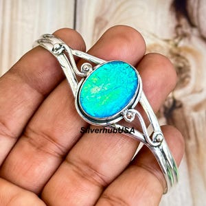 Puede incluir: Pulsera de plata con una gran piedra ovalada de ópalo azul. La pulsera tiene un diseño delicado y calado.