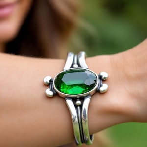 Op de afbeelding: Een zilveren manchetarmband met een grote, ovale, smaragdgroene edelsteen. De armband heeft een decoratief ontwerp met kleine, ronde accenten. De armband wordt om een pols gedragen.