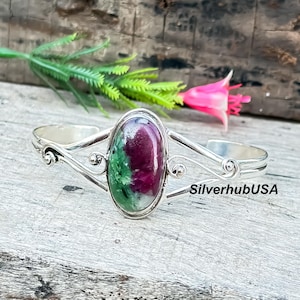 May include: A silver cuff bracelet with an oval-shaped gemstone. The gemstone features a mix of deep red and green hues. The bracelet has a decorative, open design with scrollwork and the text "SilverhubUSA".