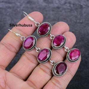 May include: A pair of silver earrings with three pink ruby stones on each earring. The earrings are dangling and have a delicate design.