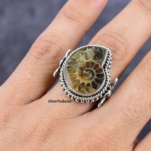 Op de afbeelding: Een ring met een gepolijst ammonietfossiel met spiraalpatroon, gezet in een zilveren montuur met een gedraaide touwrand en decoratieve zijaccenten. Het fossiel vertoont tinten bruin, beige en crème. De tekst silverhubusa is zichtbaar.