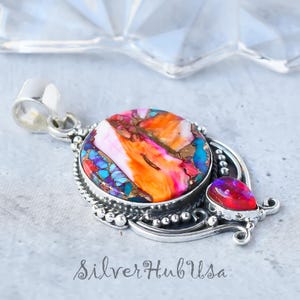 Puede incluir: Un colgante de plata con una piedra redonda grande multicolor y una piedra roja más pequeña en forma de lágrima. El colgante tiene un borde de cuentas y detalles intrincados de plata.