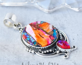 Puede incluir: Un colgante de plata con una piedra redonda grande multicolor y una piedra roja más pequeña en forma de lágrima. El colgante tiene un borde de cuentas y detalles intrincados de plata.