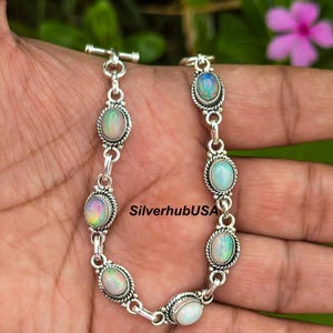 Puede incluir: Una pulsera de plata con piedras preciosas de ópalo ovaladas. Los ópalos muestran colores iridiscentes y están engastados en un bisel de plata con un borde en forma de cuerda. La pulsera está conectada por eslabones de plata y tiene un cierre de palanca.