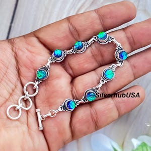 May include: Silver bracelet with iridescent blue-green stones in decorative settings. The bracelet, with a toggle clasp, is shown on a hand. The image includes the text "SilverhubUSA".