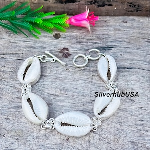 Puede incluir: Una pulsera de plata con conchas de cauri blancas. Las conchas están conectadas por eslabones de plata y un cierre de palanca. La pulsera se muestra sobre una superficie de madera desgastada, con una flor rosa y follaje verde en el fondo.