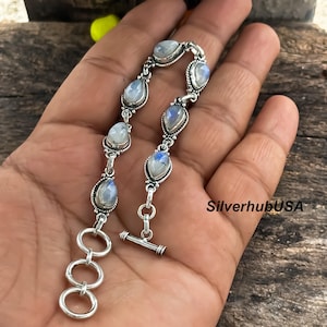 Könnte beinhalten: Ein silbernes Armband mit tropfenförmigen Mondstein-Edelsteinen. Das Armband hat einen Knebelverschluss und wird auf einer Hand vor einem Holzgrund gezeigt. Die Edelsteine haben einen blau-weißen, schillernden Glanz.