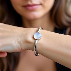 May include: A silver bangle bracelet featuring a pale blue, circular gemstone. The bracelet is displayed on a wrist, highlighting the polished metal and the smooth, round stone. The design is simple and elegant.
