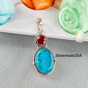 May include: A silver pendant with a large, oval, blue-green gemstone and a smaller, round, red gemstone. The pendant has a bail at the top for a chain. The text "SilverhubUSA" is visible on the right side.