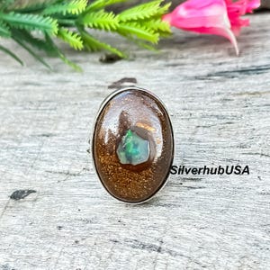 May include: Oval-shaped ring featuring a large, brown gemstone with a central, green-blue stone. The ring has a silver band and is set against a wooden surface with greenery and a pink flower in the background. The text "SilverhubUSA" is visible.