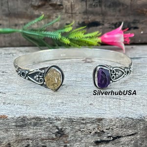 Op de afbeelding: Zilveren manchetarmband met een citrien- en amethist-edelsteen. De armband heeft een open ontwerp met sierlijke details rond de stenen. De tekst "SilverhubUSA" is zichtbaar.