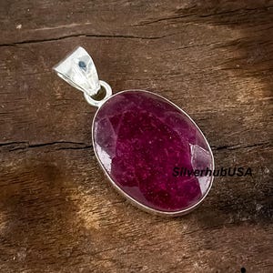 Puede incluir: Colgante de plata con una gran gema de rubí ovalada. El rubí es de un color rojo intenso y tiene una superficie facetada. El colgante tiene un diseño sencillo y elegante.