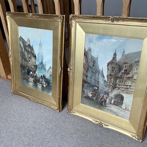 May include: Two framed landscape paintings in ornate gold frames. The artwork depicts European city scenes with buildings, people, and waterways. The paintings have a vintage aesthetic, with muted colours and detailed brushstrokes.
