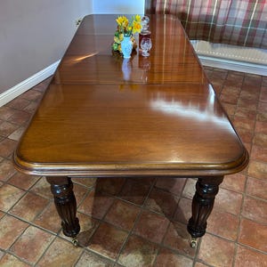 May include: A large, rectangular wooden dining table with a glossy, dark brown finish. The table has rounded edges and ornate, dark brown legs with brass casters. A small vase of flowers and decorative glassware sit on the table.