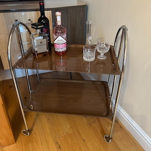 May include: A chrome bar cart with two brown wooden shelves. The cart has a bottle of red wine, a bottle of pink gin, a bottle of liqueur, a champagne flute, and a stemmed glass on the top shelf.