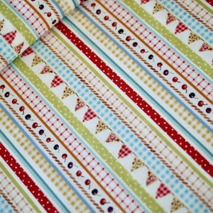 Op de afbeelding: Witte stof met rode, blauwe en groene strepen. De strepen zijn versierd met een patroon van kleine rood-wit geblokte vlaggen, bruin touw en zwarte kersen.