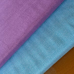Op de afbeelding: Twee stoffen stalen, één lavendelkleurig en één lichtblauw, worden diagonaal weergegeven. Beide stoffen hebben een gespikkeld patroon van kleine, donkere stippen. De stoffen zijn gevouwen en tonen hun textuur en kleurvariaties. De achtergrond is een houten oppervlak.