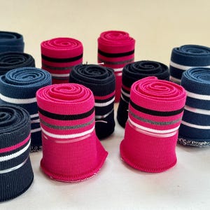 Könnte beinhalten: Gerollte Socken in verschiedenen Farben, darunter Marineblau, Pink und Schwarz, mit Streifenmustern in Weiß, Grau und Rosa. Die Socken sind ordentlich gerollt und mit einem weißen Band fixiert.