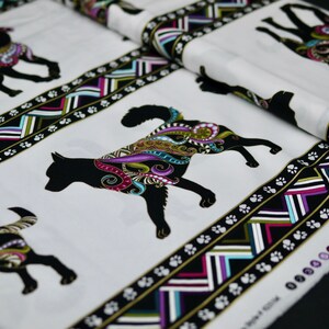 Könnte beinhalten: Weißer Stoff mit schwarzen Hundesilhouetten mit bunten Paisley-Mustern. Der Stoff hat einen Rand mit geometrischen Mustern in Schwarz, Gold, Lila und Grün sowie Pfotenabdrücken. Der Stoff ist aufgerollt.