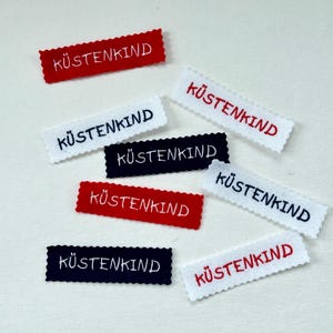 Könnte beinhalten: Eine Sammlung rechteckiger Stoffaufnäher mit dem gestickten Wort "KÜSTENKIND". Die Aufnäher sind in verschiedenen Farben erhältlich, darunter Rot, Weiß und Marineblau, mit einem gewellten Rand.
