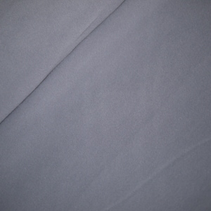 May include: A close-up shot of a folded piece of fabric. The fabric is a solid, muted gray color. The folds create diagonal lines across the frame, adding texture and dimension to the image. The fabric appears soft and smooth.