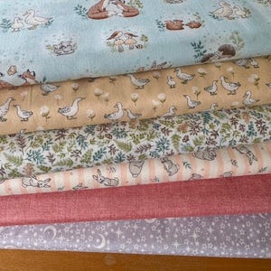Puede incluir: Una pila de telas estampadas variadas. La tela superior es azul claro con ilustraciones de animales del bosque. Debajo hay telas en color beige con gansos, estampados florales, rayas con conejos, rosa liso y una tela lavanda con estrellas y lunas.