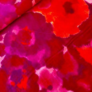 以下が含まれることがあります： 鮮やかな花柄の生地のクローズアップ。デザインは、赤、フクシア、マゼンタの色合いの大きな抽象的な花を特徴としています。生地は折りたたまれているように見え、画像全体に斜めの線ができています。