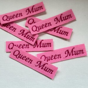 Könnte beinhalten: Rosa Filzrechtecke mit dem Schriftzug "Queen Mum" in dunklerem Rosa. Die Rechtecke sind auf einer weißen Oberfläche verstreut. Das Bild ist gut beleuchtet und der Text ist deutlich lesbar.