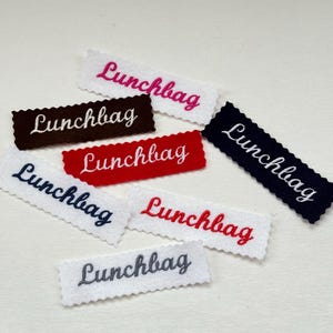 Könnte beinhalten: Eine Sammlung von bestickten Filz-Etiketten in verschiedenen Farben, jeweils mit dem Wort "Lunchbag" in Schreibschrift. Farben sind Weiß, Rot, Marineblau, Braun und Grau. Die Ränder der Etiketten haben eine dekorative, gewellte Bordüre.