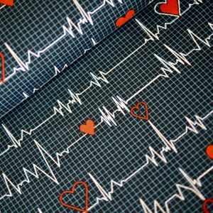 Op de afbeelding: Close-up van stof met een zwarte achtergrond en een blauw rasterpatroon. Witte lijnen die lijken op een ECG lopen over de stof, met kleine rode harten verspreid. De stof heeft een medisch of gezondheidsthema.