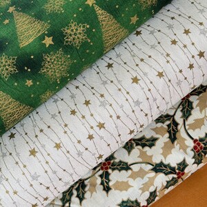 Peut inclure: Gros plan sur trois rouleaux de tissu sur le thème de Noël. Le rouleau supérieur est vert avec des sapins, des flocons de neige et des étoiles dorés. Le rouleau du milieu est blanc avec des étoiles dorées et argentées sur des lignes pointillées. Le rouleau inférieur est beige avec du houx.