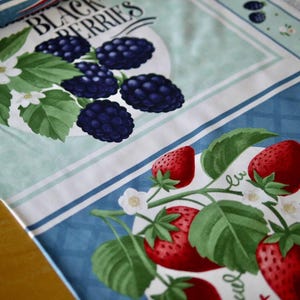 Puede incluir: Una tela blanca con un borde a rayas azul y blanco. La tela presenta un diseño de moras y fresas. El texto "Black Berries" está impreso en la tela.
