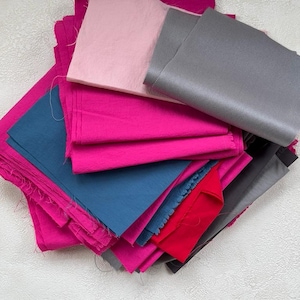 Könnte beinhalten: Eine Sammlung von Stoffmustern in verschiedenen Farben, darunter Rosa, Fuchsia, Grau, Blau und Rot. Die Stoffstücke sind gestapelt und geschichtet, wodurch verschiedene Texturen und Farbtöne zur Geltung kommen. Das Bild ist aus der Vogelperspektive aufgenommen.