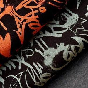 Może przedstawiać: Zbliżenie złożonej tkaniny z czarnym tłem. Tkanina ma wzór w stylu graffiti w kolorze pomarańczowym i jasnoniebieskim. Projekt zawiera abstrakcyjne kształty i litery, tworząc odważną i miejską estetykę.