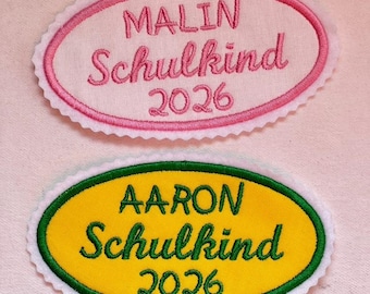 May include: Two oval-shaped embroidered patches with scalloped edges. The top patch is white with a pink border and pink text reading MALIN Schulkind 2026. The bottom patch is yellow with a green border and green text reading AARON Schulkind 2026.