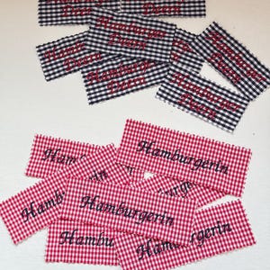 May include: A collection of fabric patches with embroidered text. The patches feature a gingham pattern in either black and white or red and white. The embroidered text reads "Hamburger Deern" or "Hamburgerin" in a cursive script. Ideal for craft projects or clothing.
