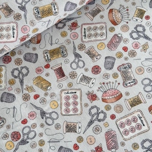Peut inclure: Tissu bleu clair avec un motif répété d'articles de couture. Le motif comprend des bobines de fil, des ciseaux, des boutons, des dés à coudre et des coussins à épingles dans des tons de marron, rose, jaune et gris. Le mot "Buttons" est visible.