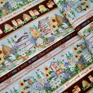 Könnte beinhalten: Baumwollstoff mit einem sich wiederholenden Muster aus Bienenstöcken, Sonnenblumen, Lavendel und Gänseblümchen. Das Design enthält Texte wie Bee Kind, Bee Honest und Bee Positive sowie Honiggläser und Bienen auf hellblauen und braunen Streifen.
