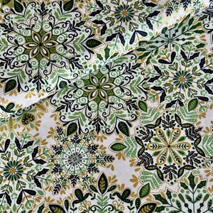 May include: Close-up of a white fabric featuring a repeating pattern of green, black, and gold floral and leaf designs. The fabric has a festive, holiday theme, with a snowflake-like appearance.