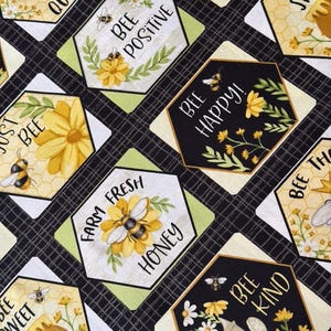 May include: Black and white fabric with a repeating pattern of hexagonal shapes. Each hexagon features a bee and a floral design with a motivational quote. The quotes include "Bee Positive", "Bee Happy", "Bee Kind", "Bee Sweet", "Farm Fresh Honey", "Just Bee", and "Bee The Queen".