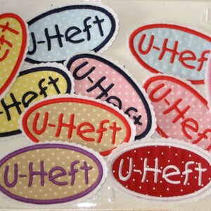 Könnte beinhalten: Eine Sammlung von acht ovalen Stoff-Patches mit weißen Punkten. Die Patches sind in verschiedenen Farben, darunter Rot, Rosa, Gelb, Blau und Beige. Jeder Patch hat den Text "U-Heft" in weißer, roter oder lila Schrift.