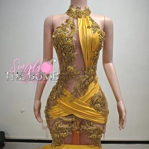 May include: A yellow halter-neck dress with gold embellishments and a sheer panel. The dress features intricate gold embroidery and a mermaid-style skirt. The dress is displayed on a mannequin.