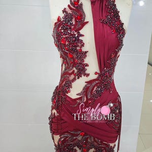 May include: A burgundy halter-neck dress with intricate red beading and embellishments. The dress features a fitted bodice, a sheer panel, and a draped skirt. The text "Simply The Bomb" is visible.